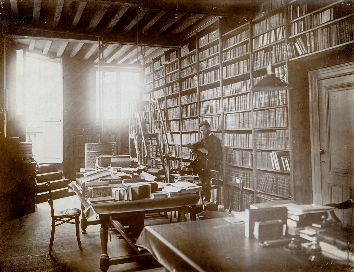 Emmanuel de Bom, lezend en leunend tegen de rekken. In zijn bureau in de Stadsbibliotheek., ca. 1906