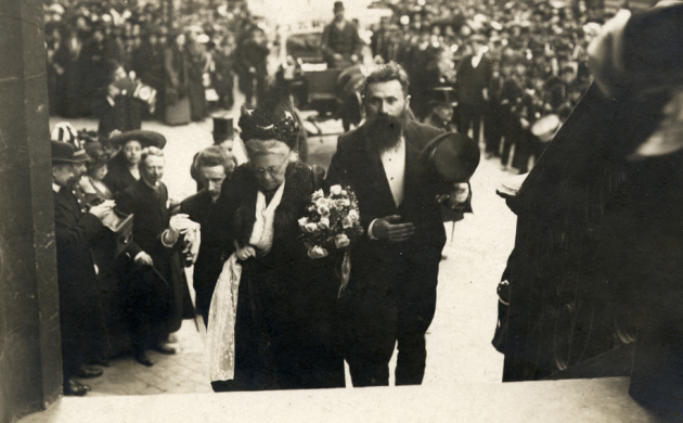 Zwartwitfoto van Virginie Loveling en René de Clercq in feestelijke kledij die de trappen oplopen aan de ingang van de huldebetoging