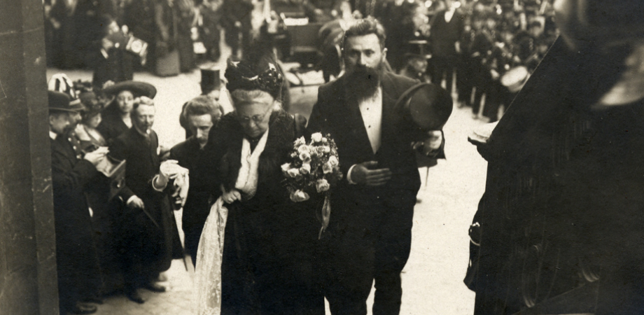 Zwartwitfoto van Virginie Loveling en René de Clercq in feestelijke kledij die de trappen oplopen aan de ingang van de huldebetoging